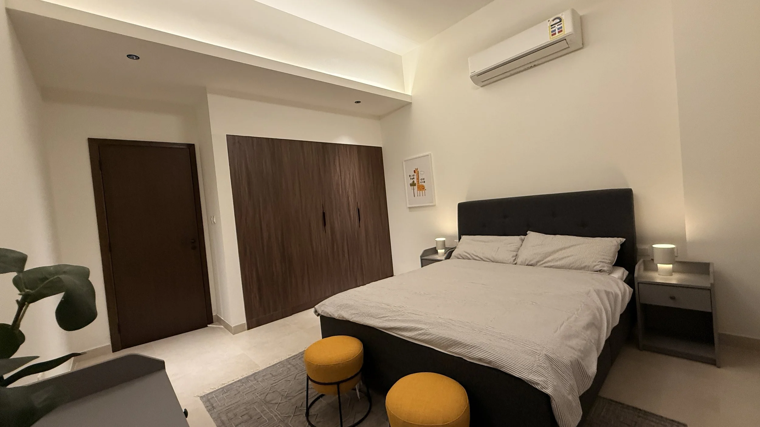 Modern bedroom with a dark upholstered bed, light bedding, two matching nightstands with lamps, a wooden closet, a framed artwork on the wall, air conditioning unit, .residences oman, freehold, apartments visa residency muscat residences in duqm