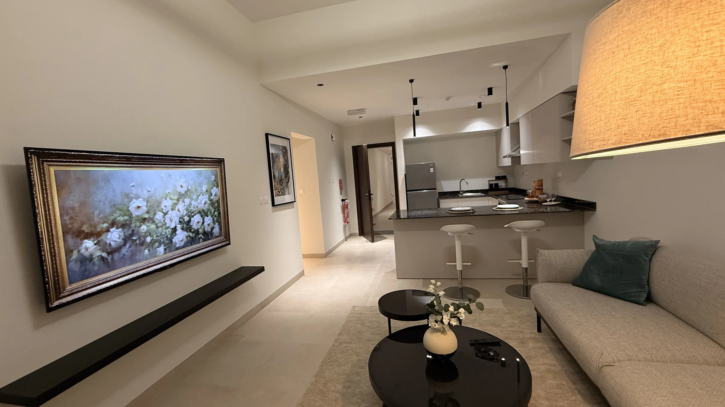 Modern living room with paintings on the wall, a beige sofa with a dark green pillow, a round black coffee table with a vase of flowers, an open kitchen .residences oman, freehold, apartments visa residency muscat residences in duqm