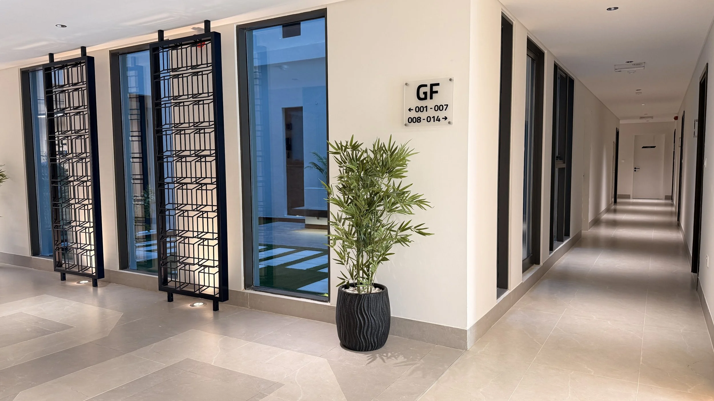 Modern corridor with beige tile flooring, white walls, and black-framed windows and decorative panels. A potted plant is near the wall with a sign indicating room numbers.