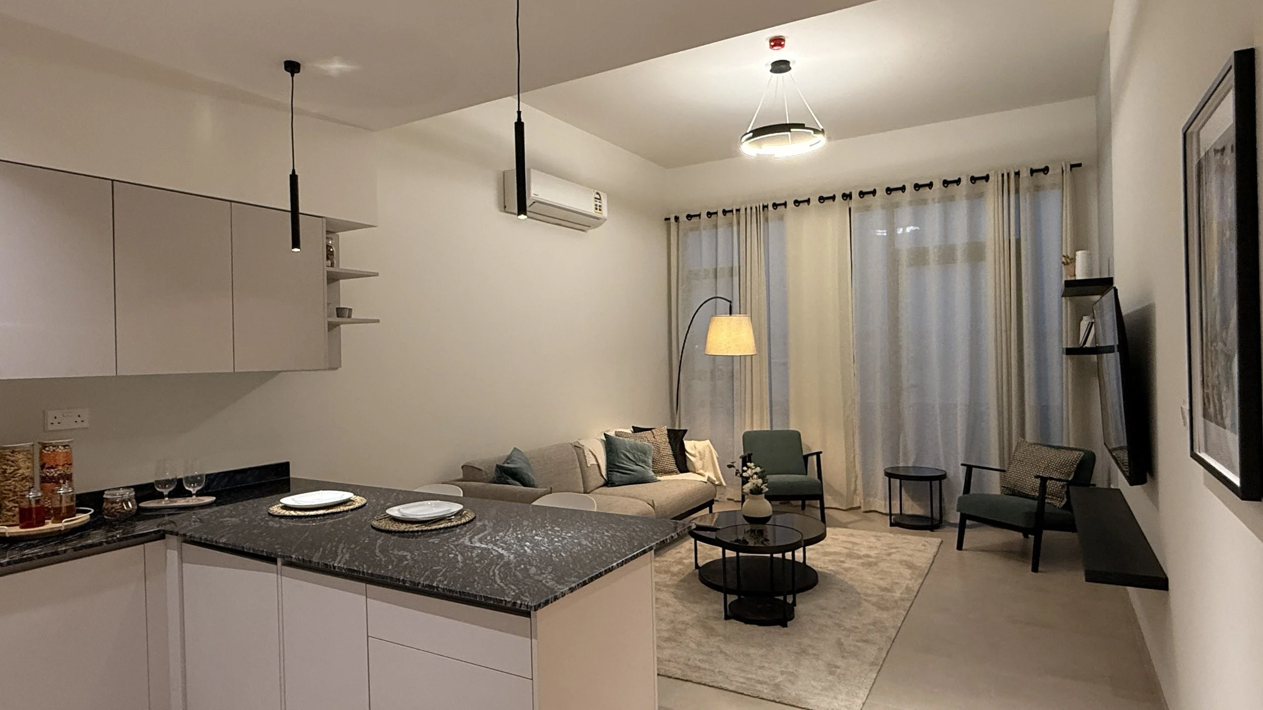 Living room with a beige sofa, green armchair, and a round coffee table, with curtains covering large windows, and a kitchen counter w.residences oman, freehold, apartments visa residency muscat residences in duqm