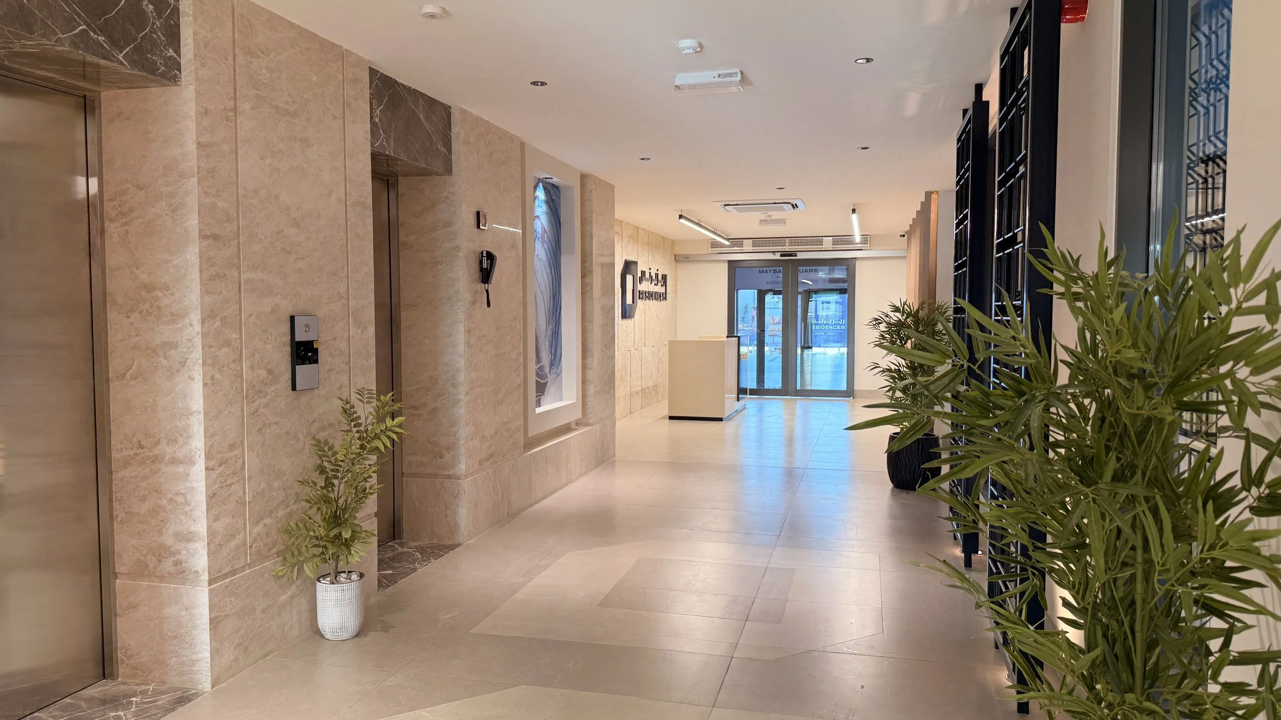 Modern hotel or office lobby with beige marble walls and tiled flooring, potted plants, glass doors in the background, and black metal decorative elements.