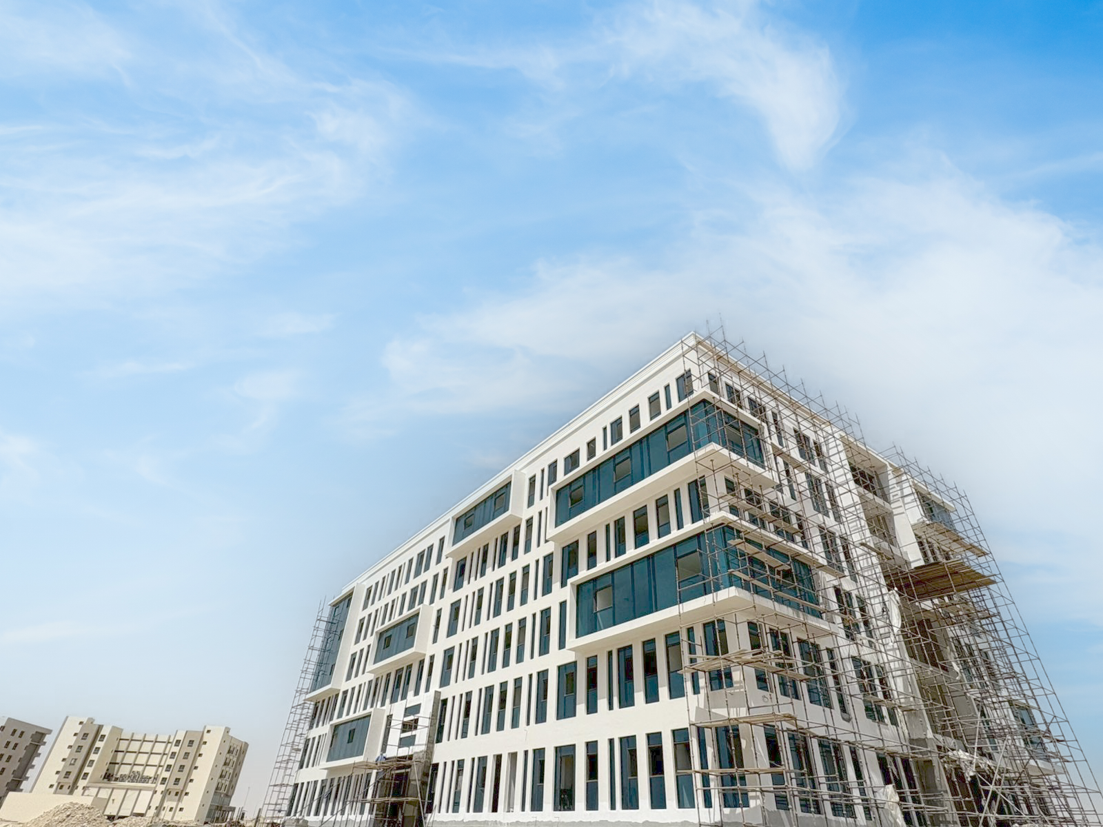 Modern white Residences Oman Duqm multi-story building under construction with scaffolding, blue sky, and scattered clouds in the background.