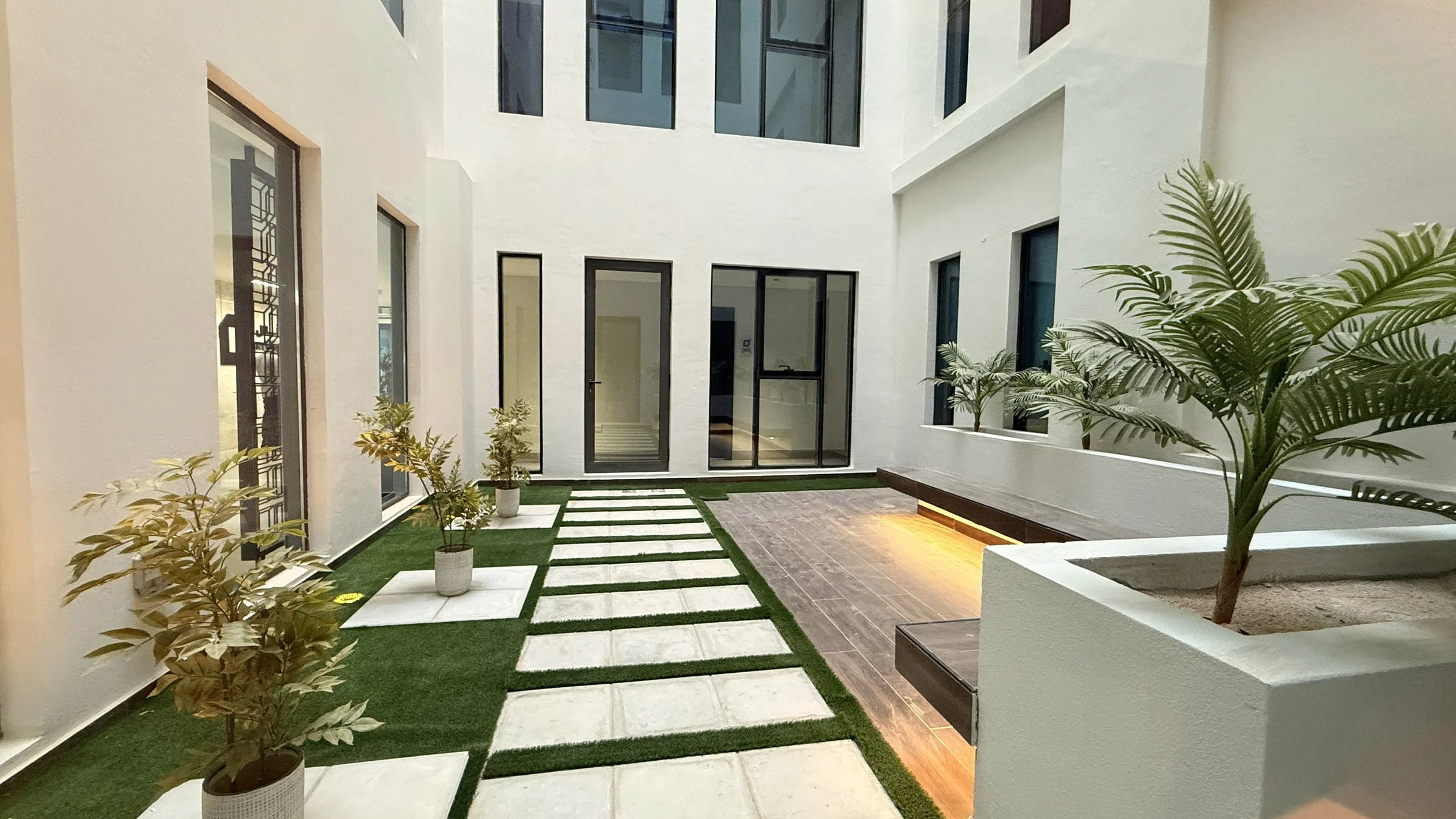 Interior courtyard with white walls, large glass doors and windows, potted plants, artificial grass, and wooden flooring, illuminated by warm lighting.