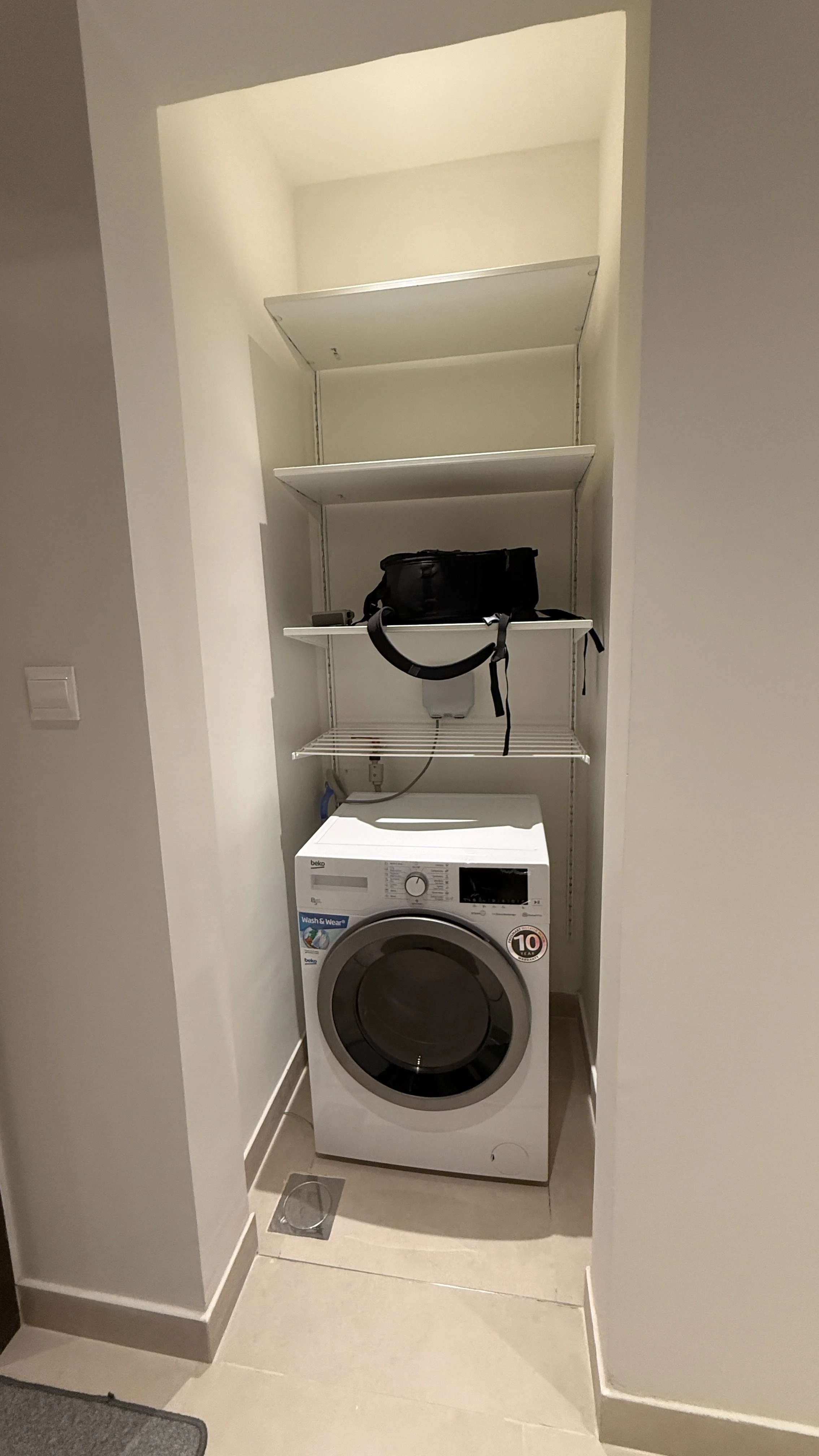 A laundry area with a front-loading washing machine beneath open shelving, a black bag and other items on the shelves, and a small vent on the floor..residences oman, freehold, apartments visa residency muscat residences in duqm