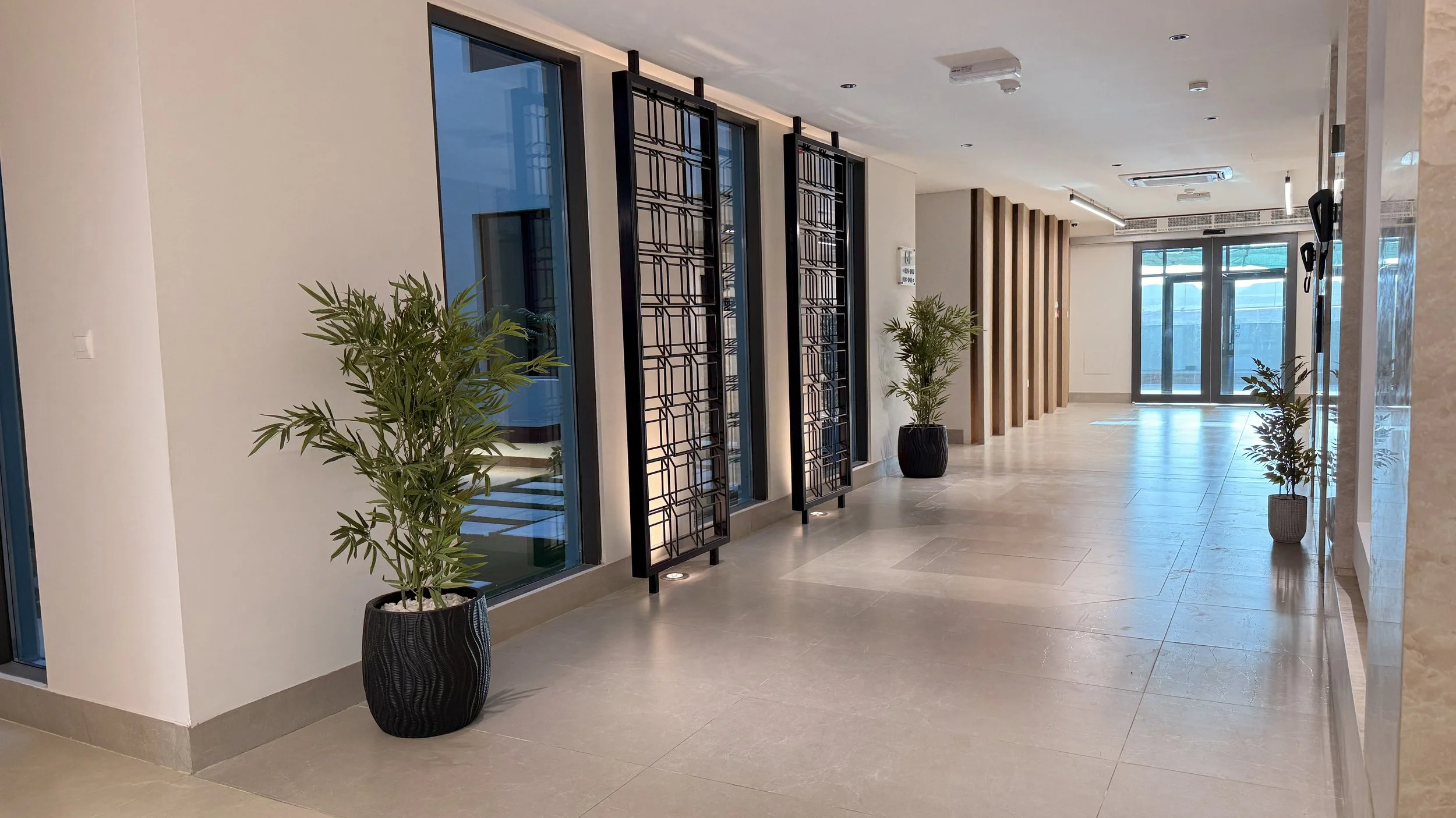 Modern indoor lobby with beige tiled floor, potted plants, decorative black screens, glass doors, and a wooden slat wall near the ceiling.