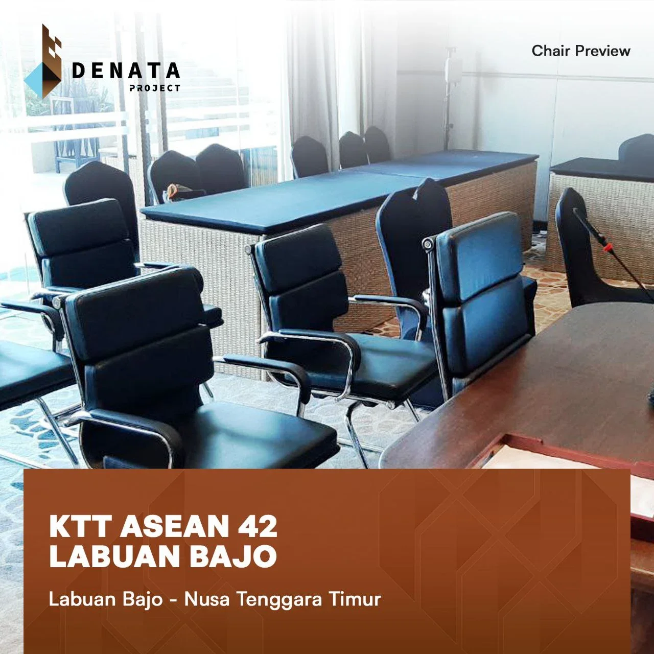 Conference room with black leather chairs around a wooden table, a long table with a blue surface at the back, and a projector or microphone stand. There are large windows with curtains and a logo and text overlay in the upper left corner and bottom 