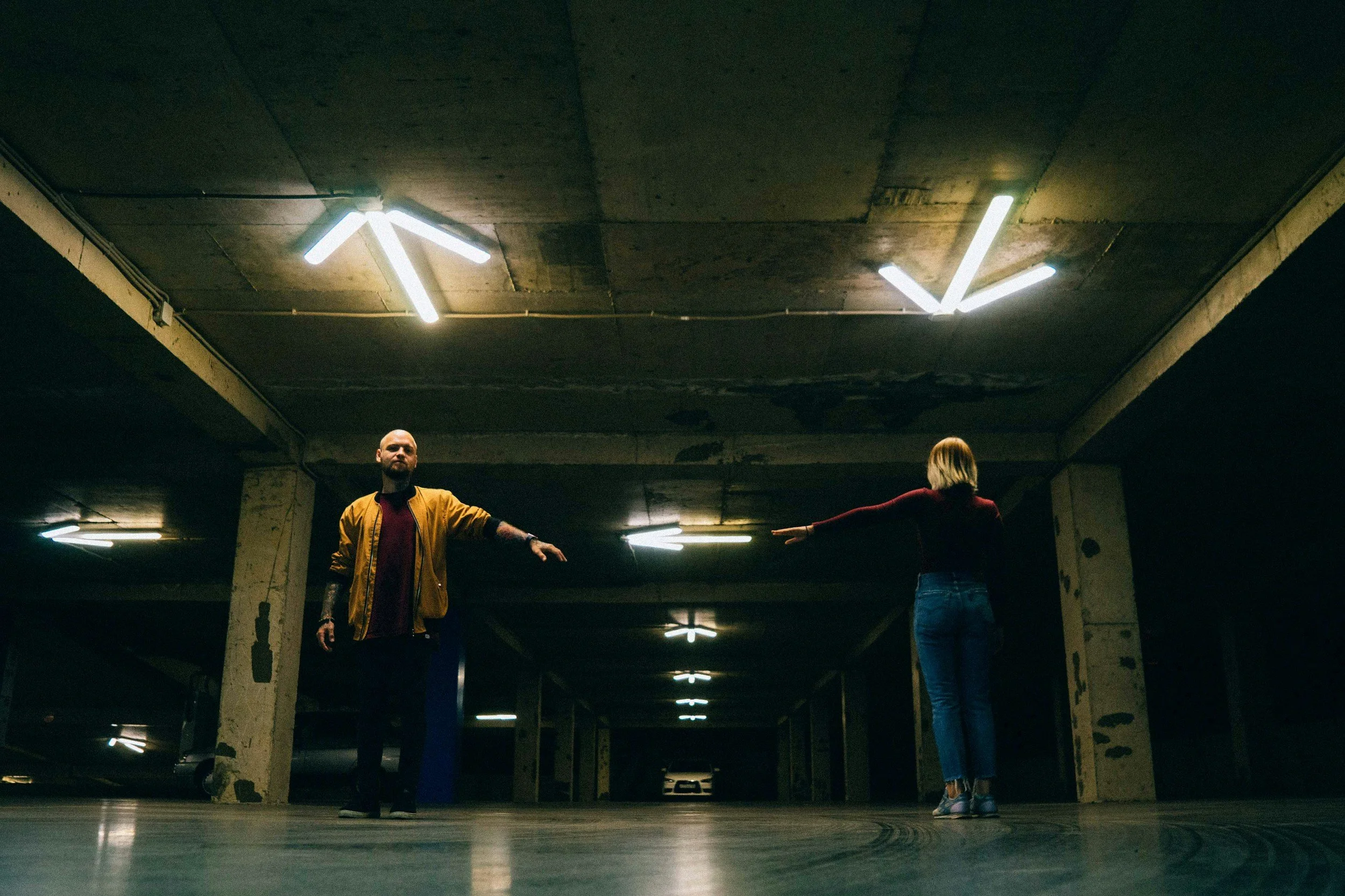 Two people standing apart in a parking garage with their arms extended toward the space between them.