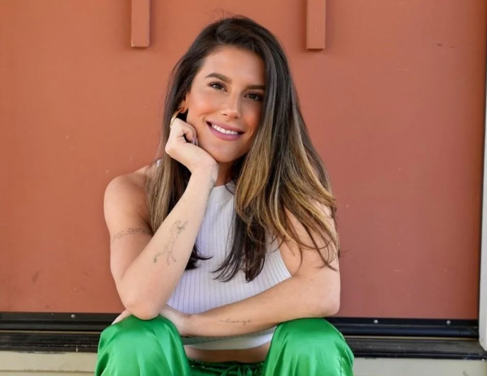 A woman sitting and smiling, with long brown hair, wearing a white sleeveless top and green pants, against a reddish-brown wall.