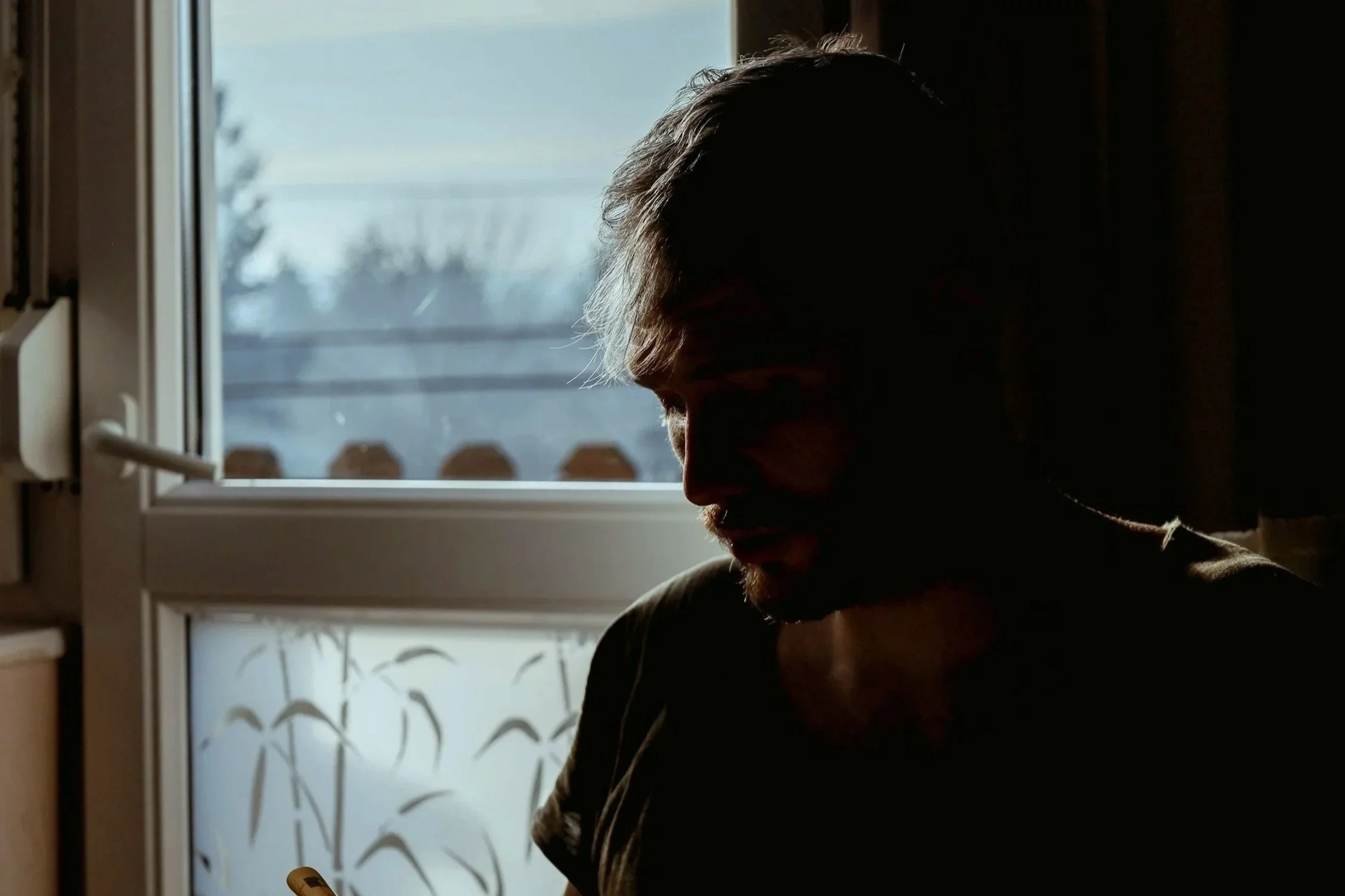 Man sitting beside a window with his head lowered in a dimly lit room.