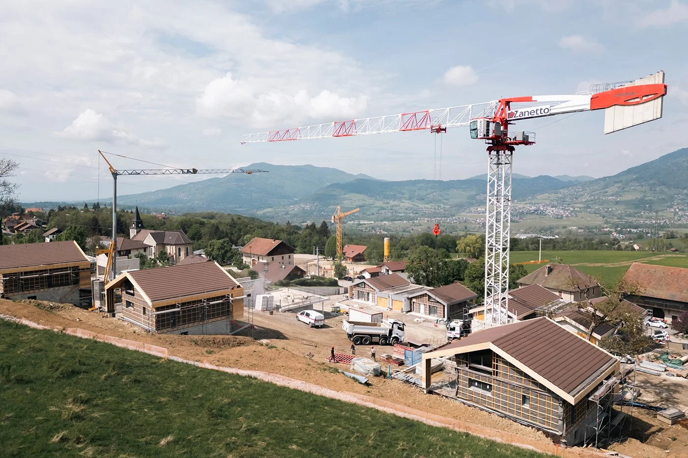 Chantier de construction avec une grue rouge et blanche. Plusieurs structures en béton en cours de construction, maisons et paysages ruraux en arrière-plan.