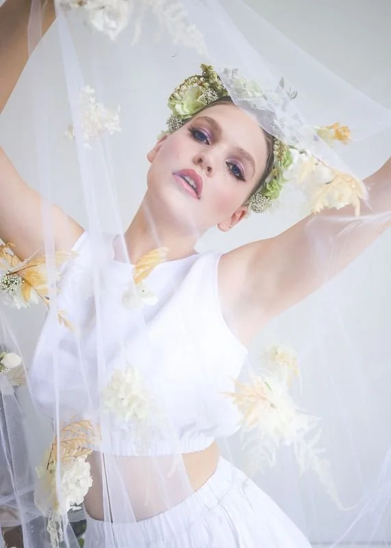 Woman dressed as a bride, wearing a white dress and floral headpiece, surrounded by white and yellow flowers and veil.
