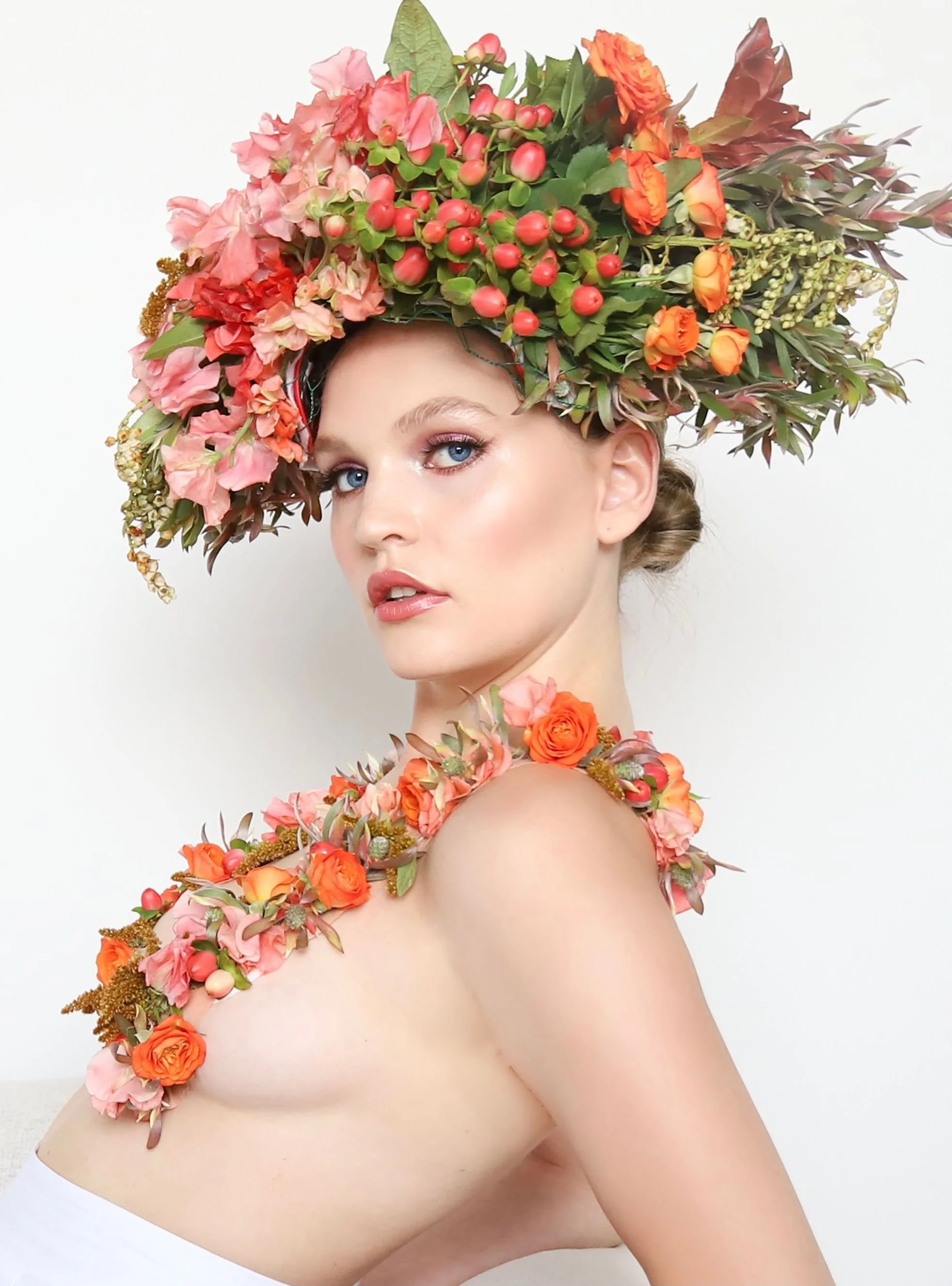 A woman wearing a large floral headpiece and matching floral shoulder accessory, both made of pink and orange flowers with green leaves, against a white background.