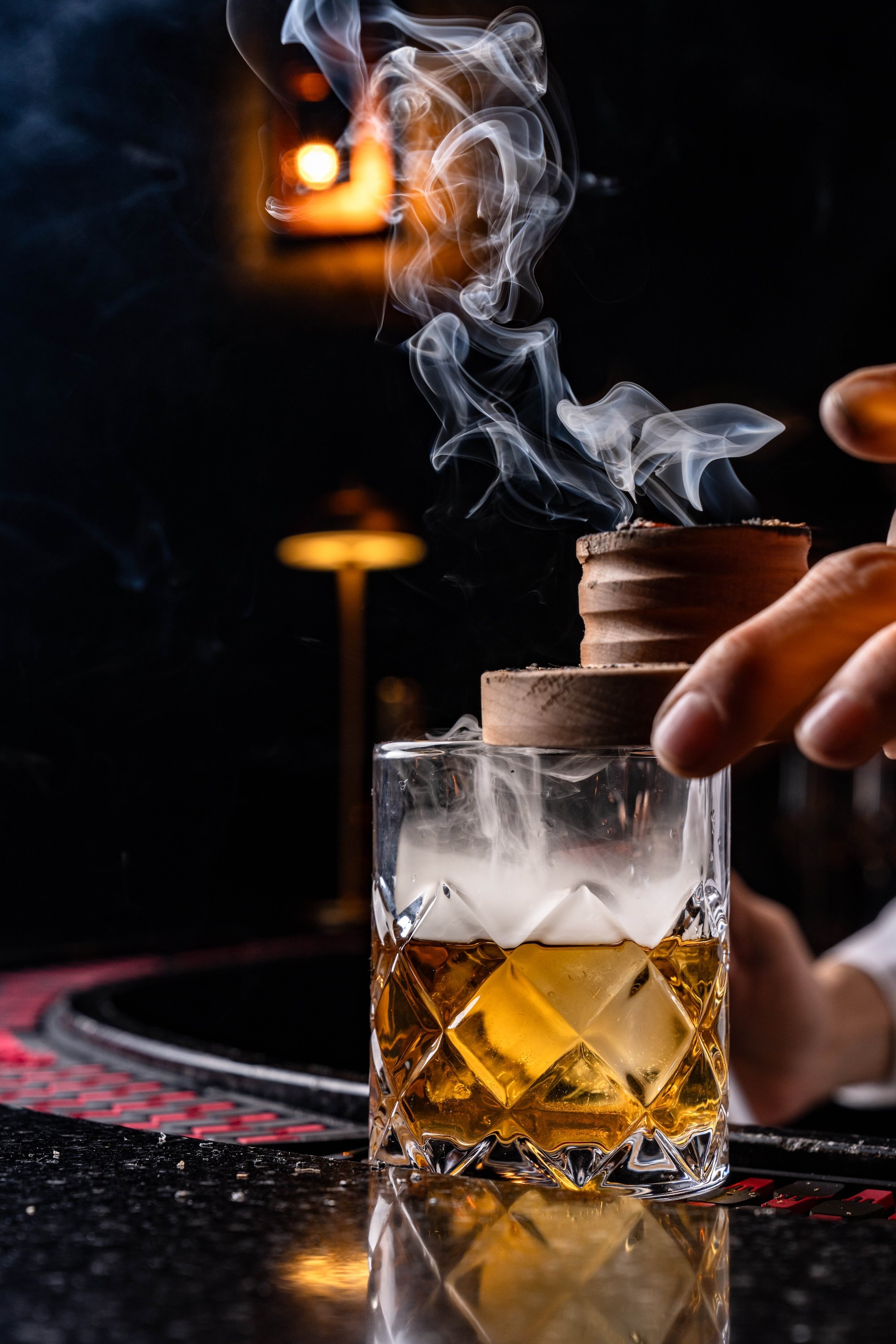 A hand holding a cocktail glass with a gold-colored drink, topped with a layer of foam, while smoke or vapor rises from a wooden container on a dark bar surface background.