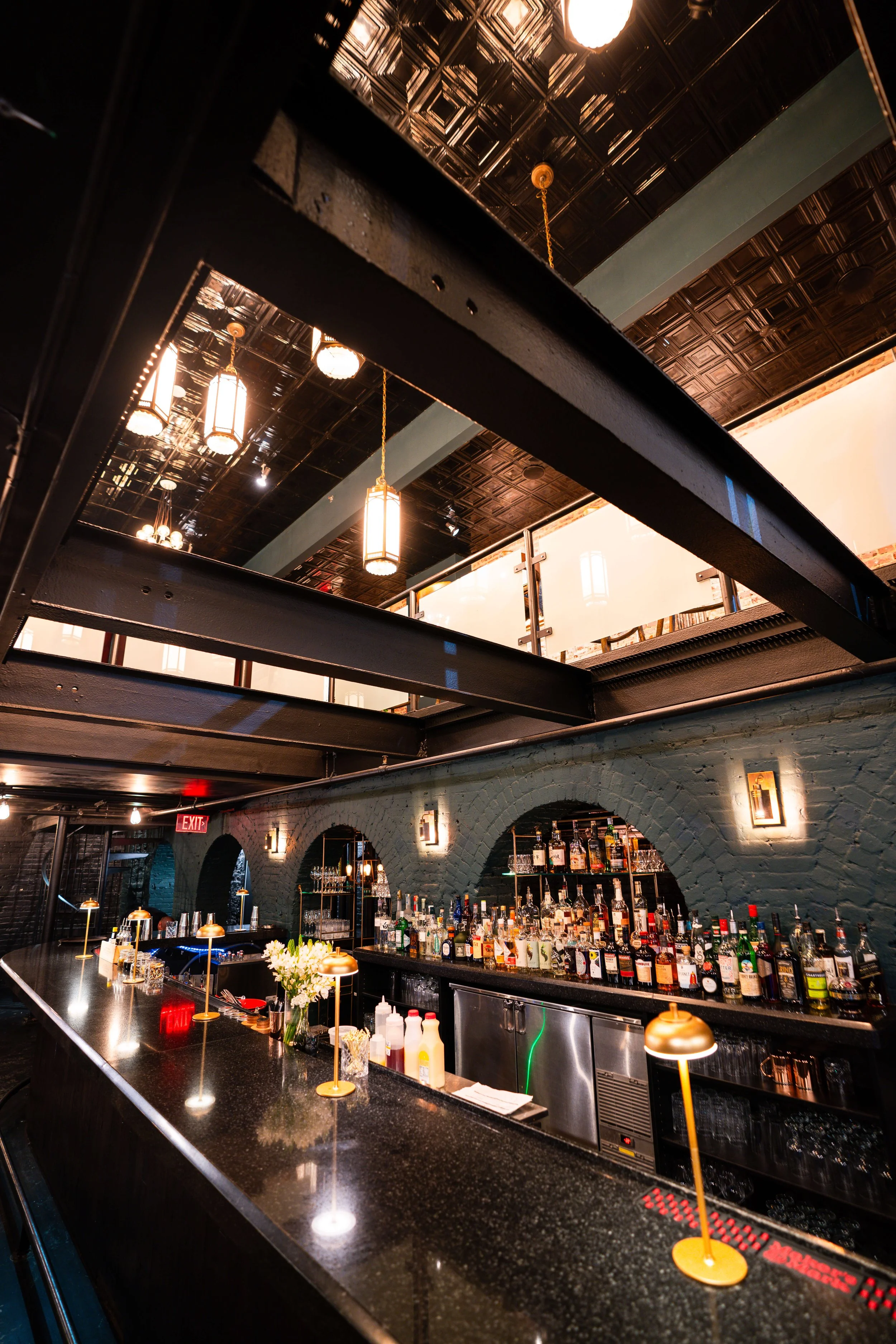 A bar in a restaurant with a dark granite countertop, a variety of liquor bottles on shelves, and modern hanging pendant lights with warm glow. Exposed brick wall with arches and wall sconces, and a ceiling with metallic finish.