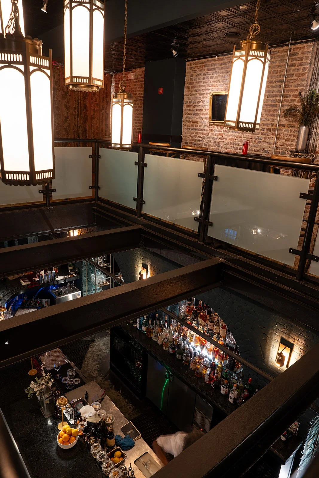 Interior of a bar with hanging lantern-style lights, exposed brick walls, and a glass railing. The bar counter is filled with lemons, glassware, and bottles of alcohol. The lower level shows more alcohol bottles and bar equipment.