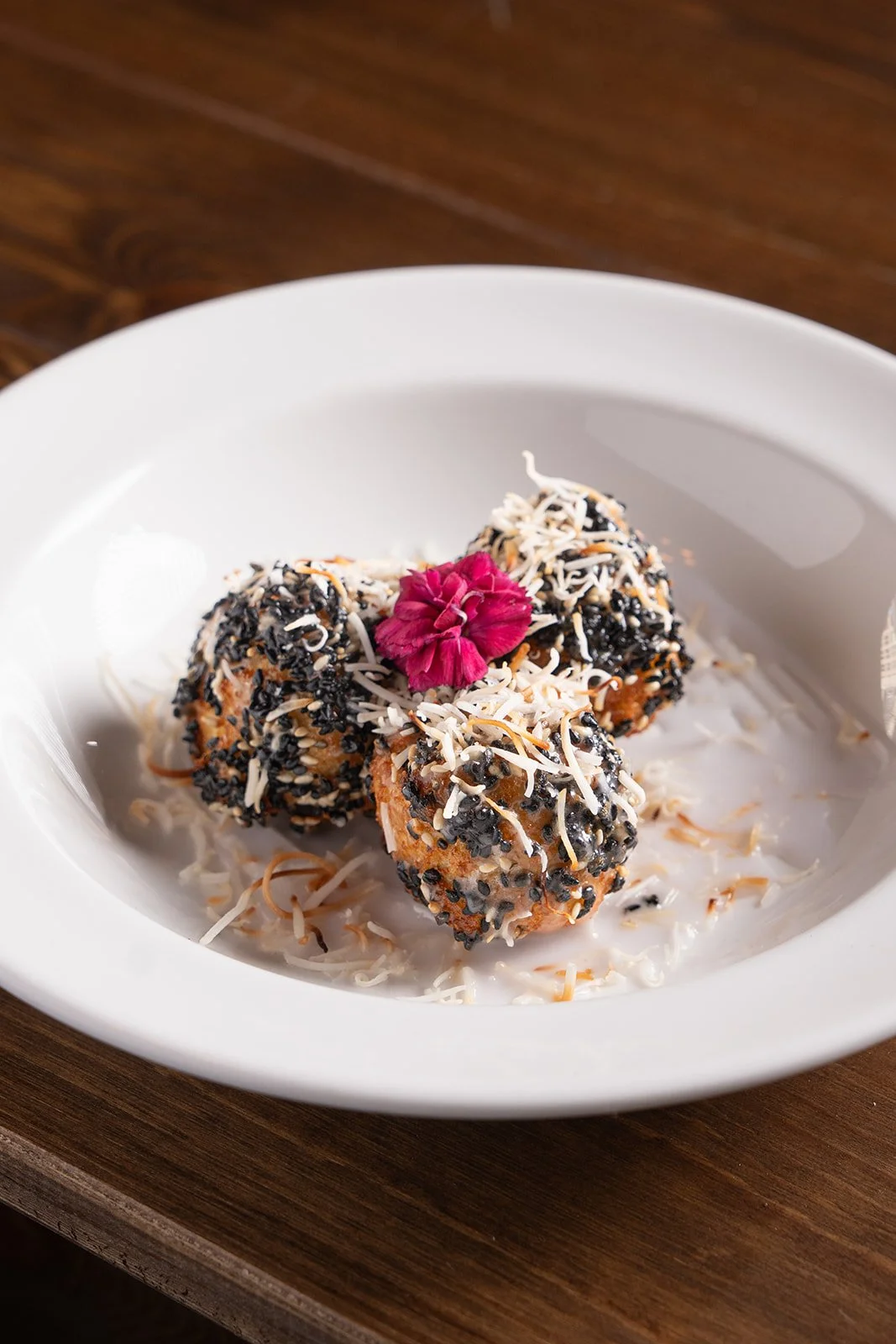 Three round food balls covered in black and white sesame seeds and shredded cheese, garnished with a pink flower on top, served on a white plate with coconut flakes and sauce.