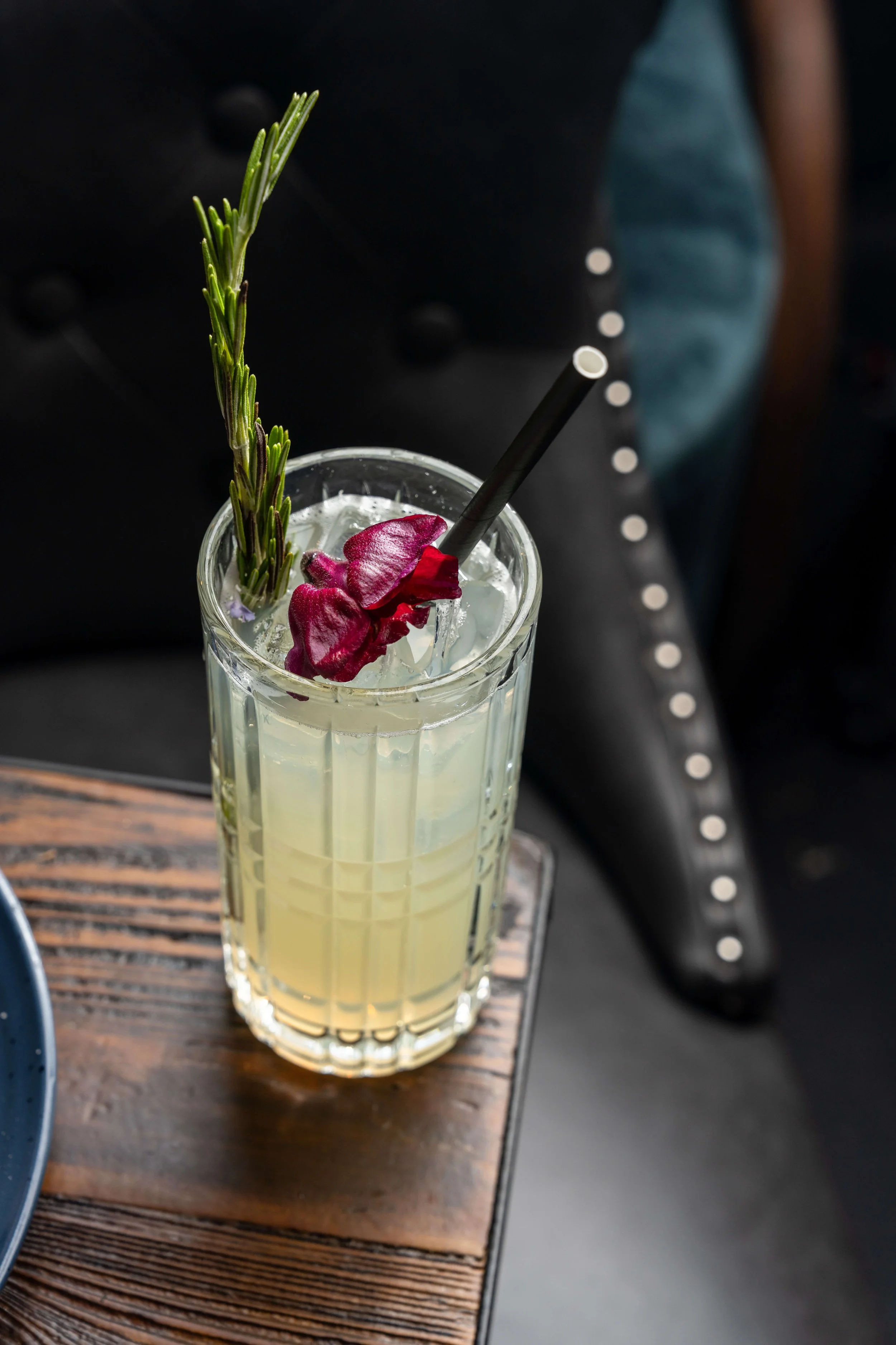 A tall glass of a pale yellow beverage with ice, garnished with a sprig of rosemary and a single edible flower petal, with a black straw, placed on a wooden table.
