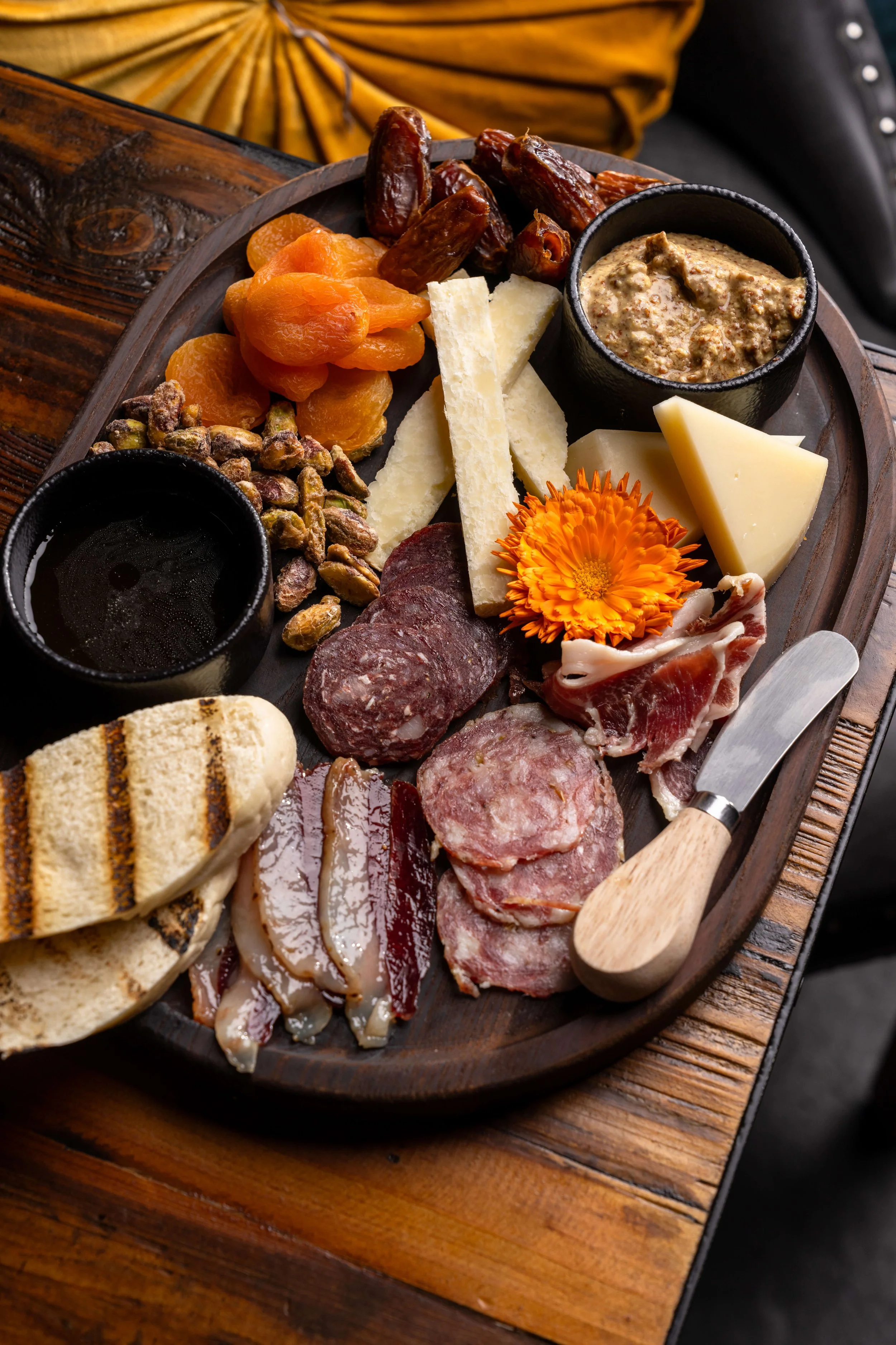Charcuterie board with dried apricots, dates, nuts, cheeses, cured meats, and a small container of mustard or dip, decorated with an orange flower, on a wooden table.