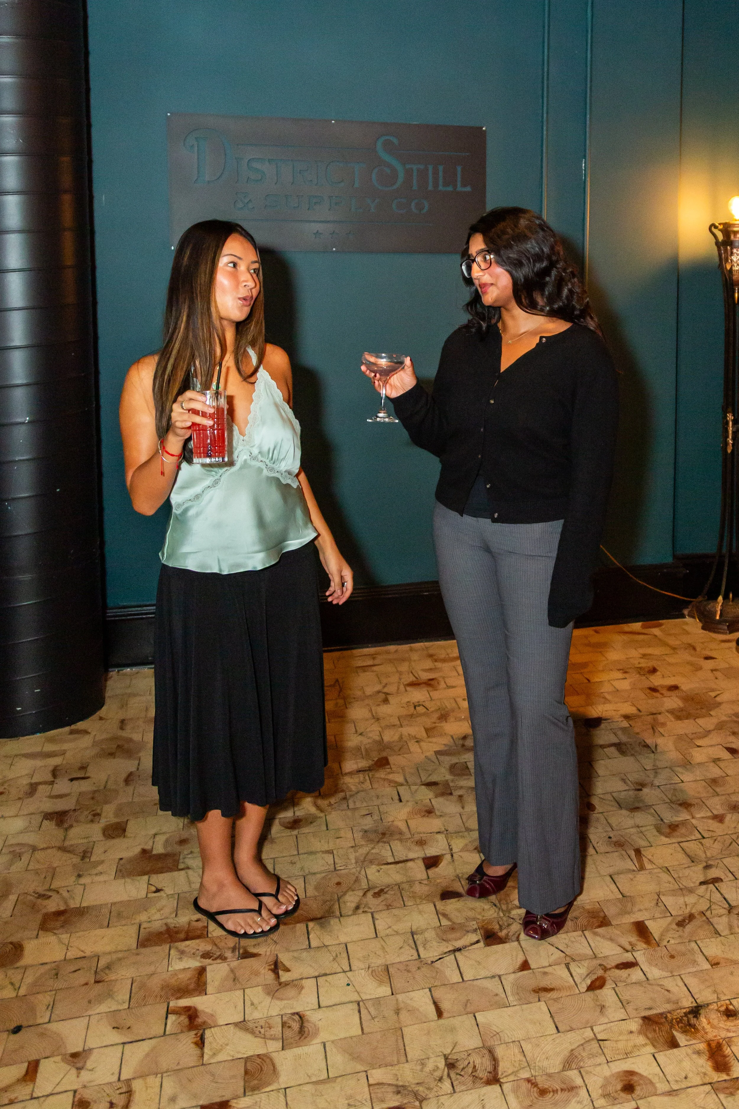 Two women standing and conversing at an indoor event, holding glasses of drinks. One woman is dressed in a satin camisole and long skirt, while the other wears a black blazer and gray pinstripe pants. They are in front of a teal wall with a sign reading 'District Still & Supply Co.'