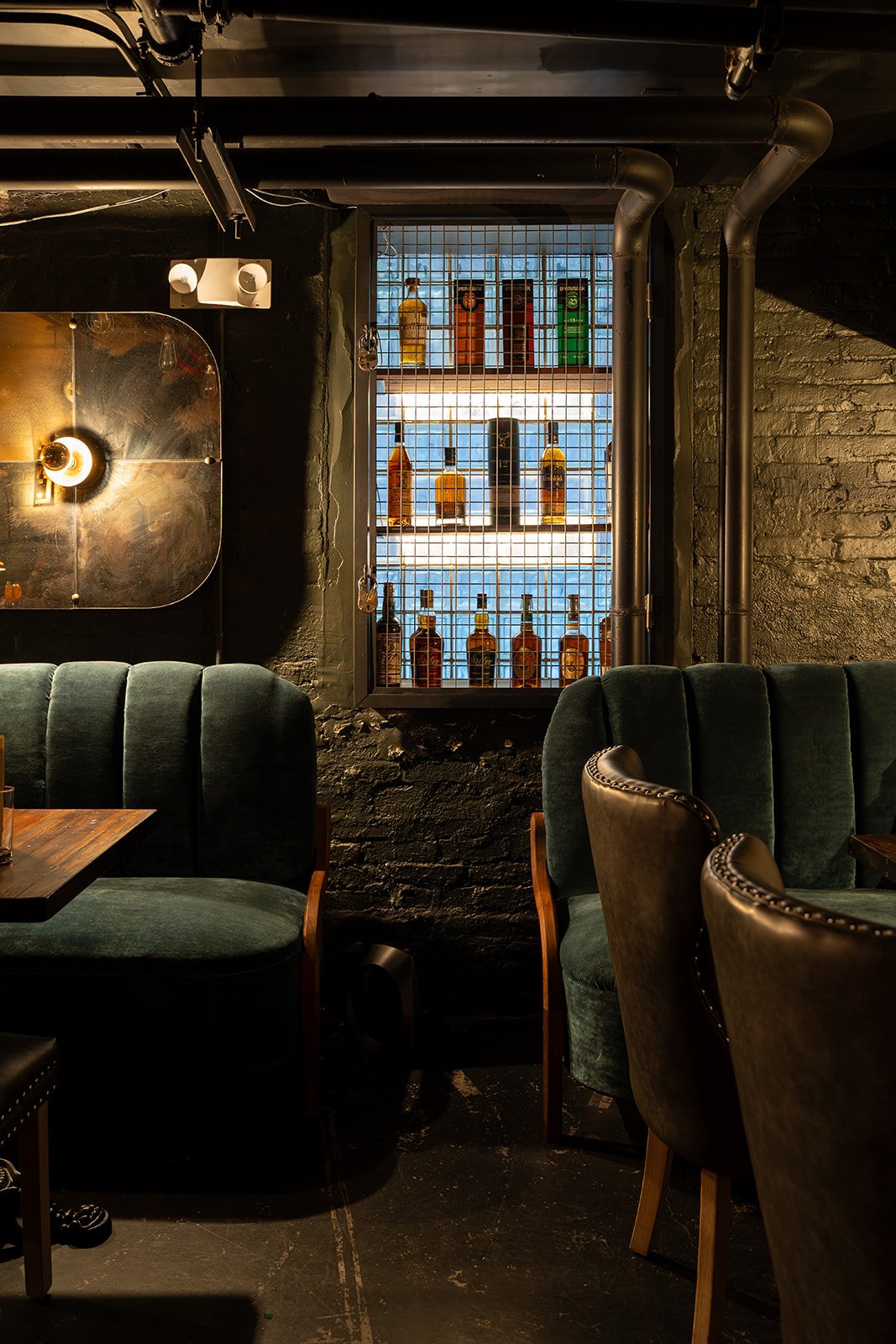 A dimly lit bar with a window showcasing shelves of brown and green liquor bottles behind a wire mesh. Dark green plush chairs and a wooden table are in the foreground, with black brick walls and exposed pipes.