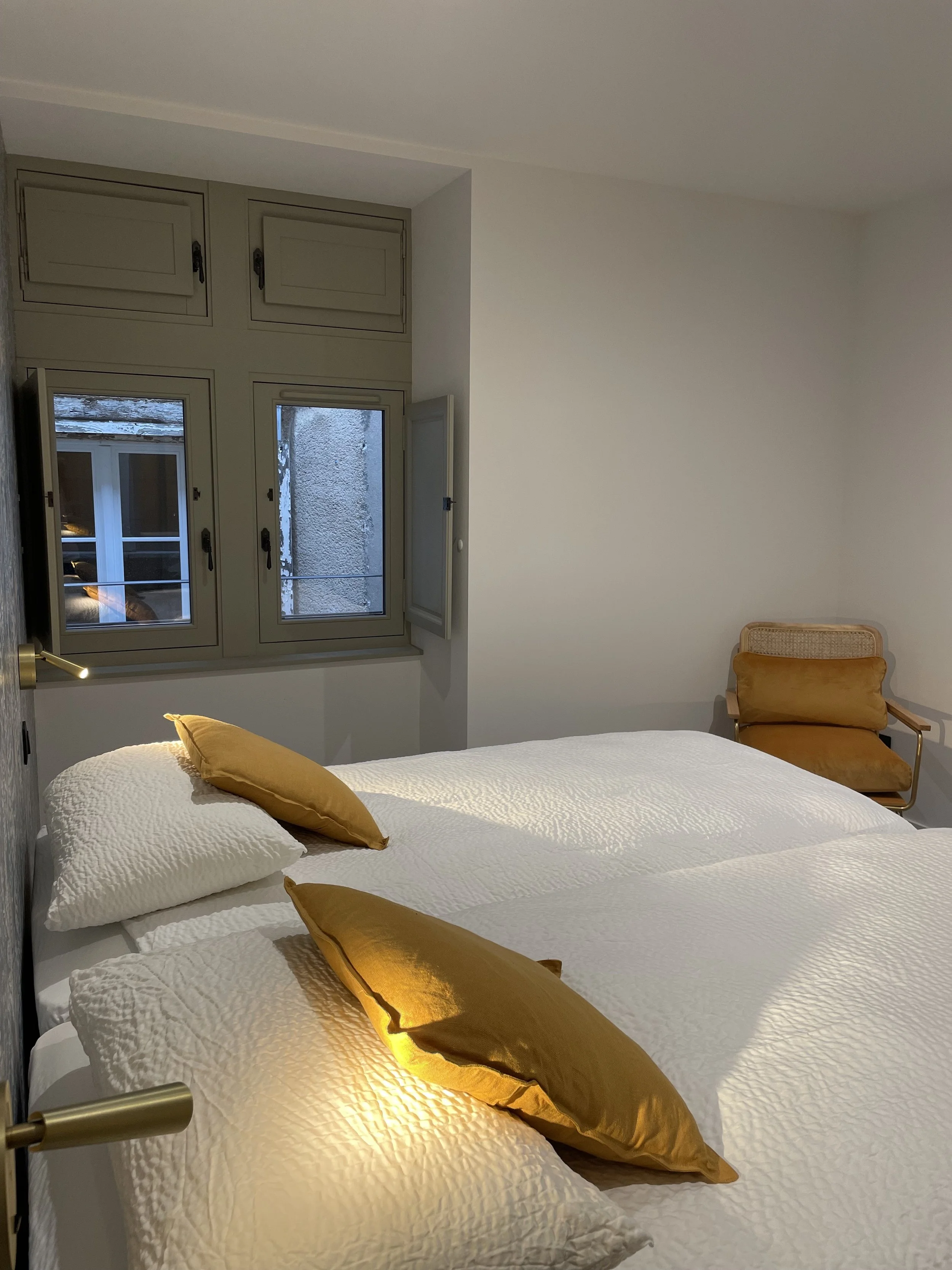 Chambre avec deux lits blancs, trois oreillers dorés, une chaise en rotin avec coussin doré, fenêtres avec volets fermés.