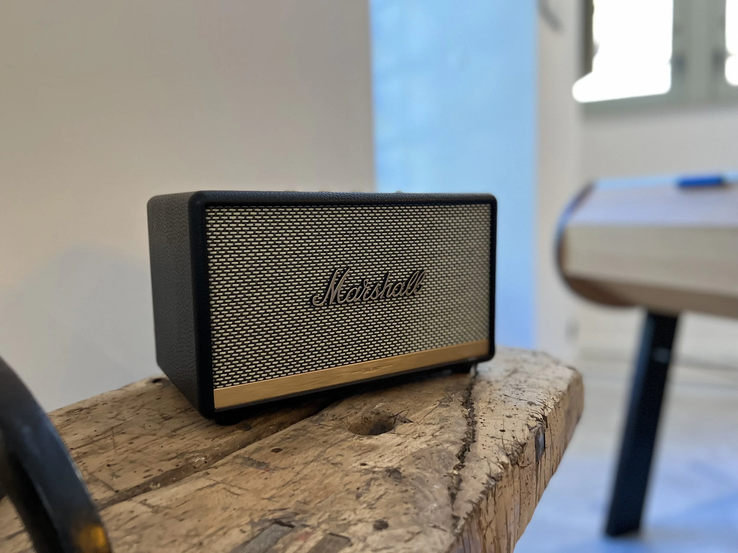 Enceinte Marshall noire avec une grille métallique, posée sur une table en bois brut, dans une pièce lumineuse.