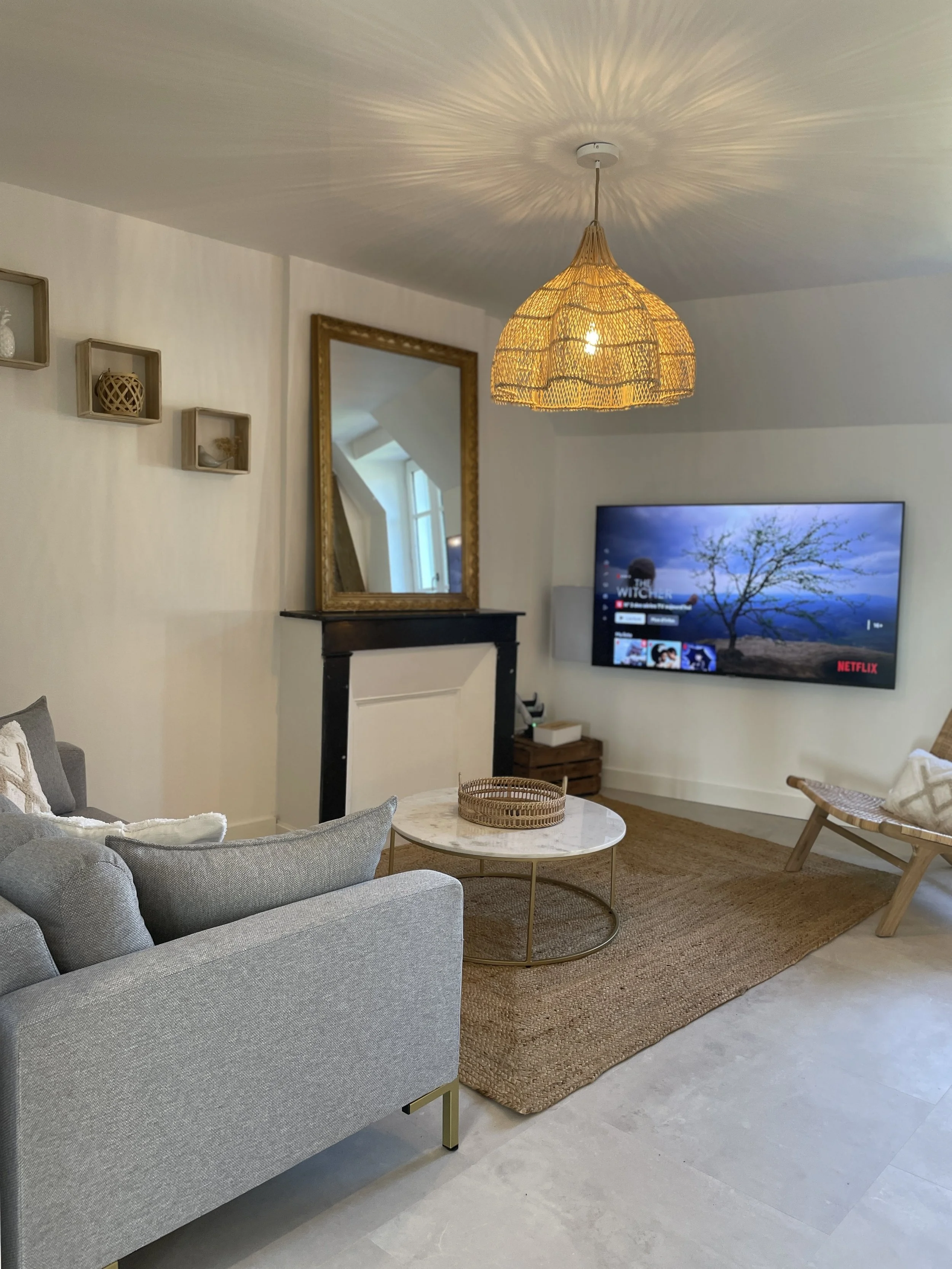 Salon moderne avec canapé gris, table basse en marbre, tapis en fibre naturelle, mur blanc, grand miroir avec cadre doré, suspension en rotin, télévision à écran plat affichant Netflix, paravent en bois, décorations en panier et céramique.
