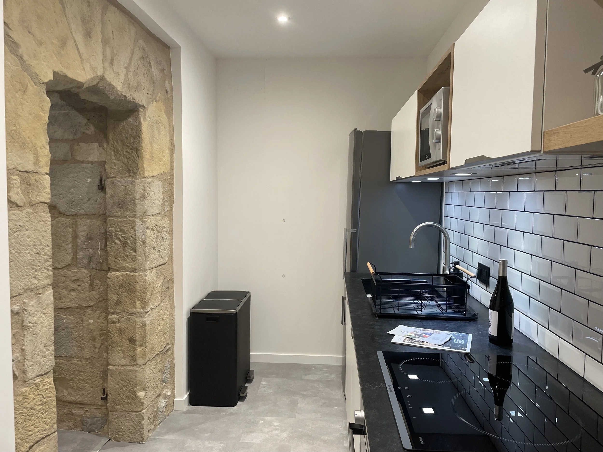 Cuisine moderne avec mur en briques blanches, plan de travail noir, évier, micro-ondes encastré, réfrigérateur gris et un mur en pierre beige. Il y a également une bouteille de vin et des brochures sur le plan de travail.