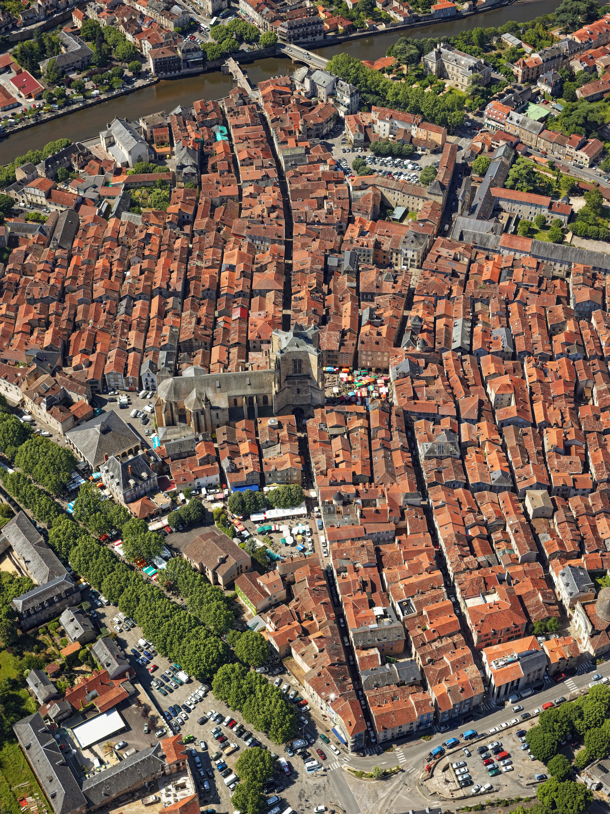 Vue aérienne d'une vieille ville avec des toits en tuiles rouges, une église ancienne au centre, et un fleuve à l'arrière-plan.