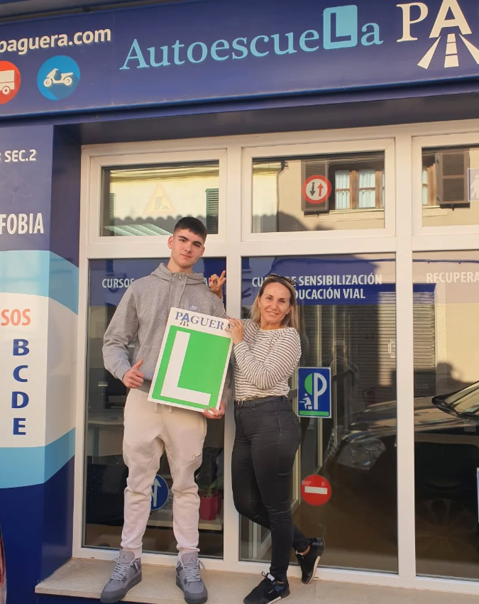 Joven y mujer posando con señal de conductor principiante en frente de una escuela de manejo.