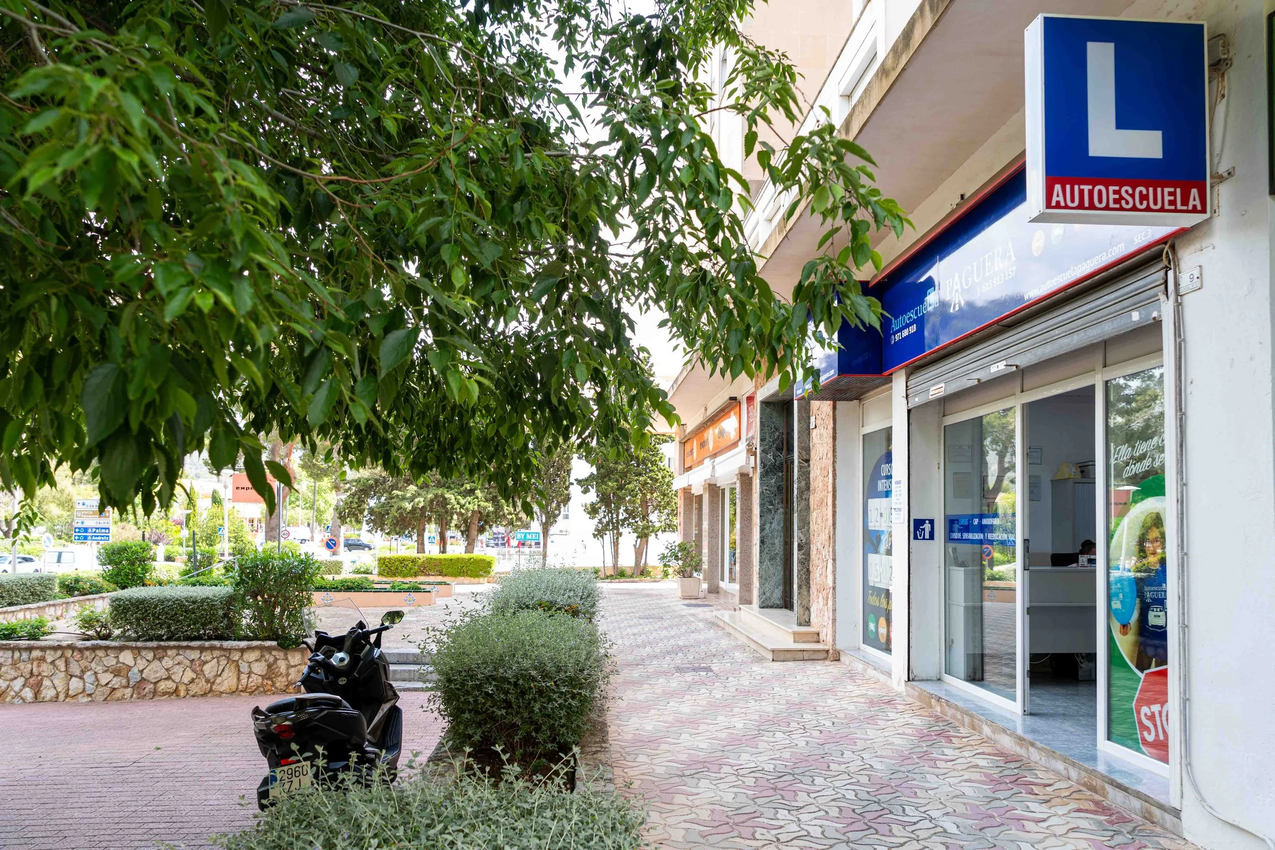 Vista de una acera con árboles y arbustos, un coche de motocicleta, y varias tiendas con letreros azules y una señal de autoescuela con una L y la palabra 'AUTOESCUELA' en letras rojas.
