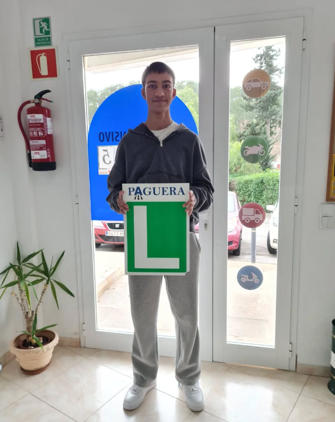 Joven de pie en un interior, sosteniendo señal de instructor de manejo con la letra 'L' y la palabra 'PAGUERA', frente a puerta de vidrio con símbolos de diferentes vehículos, plantas y señal de salida de emergencia.