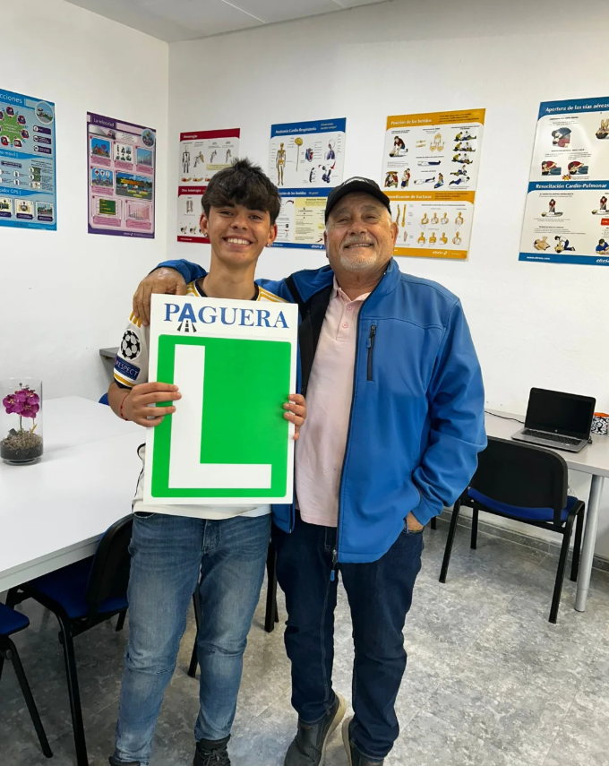 Joven y hombre en una sala con posters de primeros auxilios, uno sostiene un cartel con la letra L y la palabra "PAGUERA".