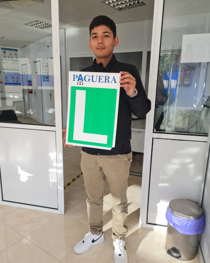 Joven sosteniendo cartel de agujero deInstructor de auto con la letra L en verde y la palabra 'PAGUERA' en azul en una oficina.