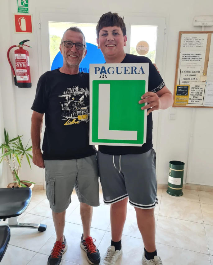 Dos hombres, uno mayor y otro joven, posando juntos en una oficina de inmigración; el joven sostiene un cartel que dice 'PAGUERA' y tiene un gran signo de interrogación verde.