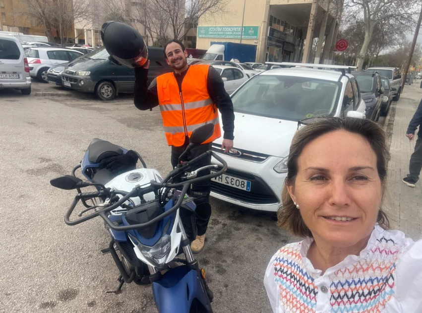 Dos personas en una calle con autos aparcados, una mujer en primer plano y un hombre en el fondo. La mujer está sonriendo y usando una blusa blanca con detalles coloridos, el hombre lleva un chaleco de seguridad naranja y sostiene un casco en la mano