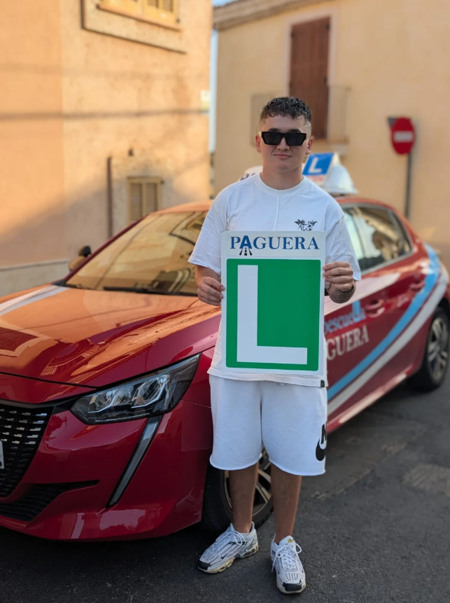 Joven con gafas oscuras, camiseta blanca, shorts blancos y tenis blancos, sostiene un cartel con una ‘L’ grande en verde y la palabra ‘Paguera’ en la parte superior, de pie frente a un coche de autoescuela rojo con señal de ‘L’ en el techo y señal de