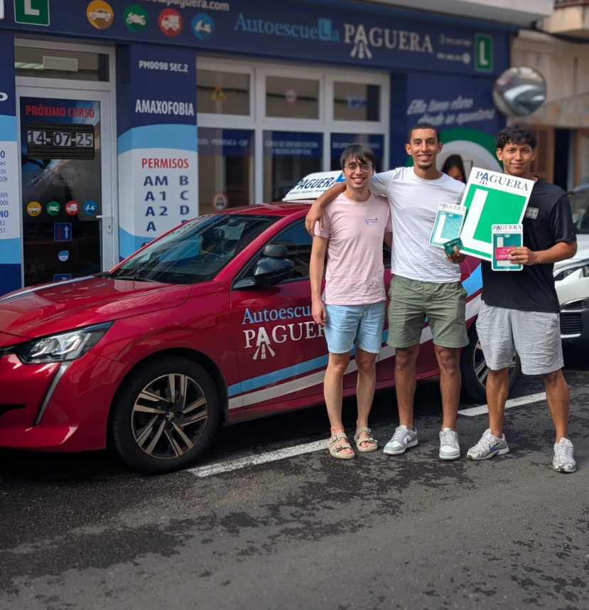 Tres personas celebrando frente a una academia de conducir, con un coche rojo y carteles que indican que han aprobado. La academia se llama Autoescuela Paguera.