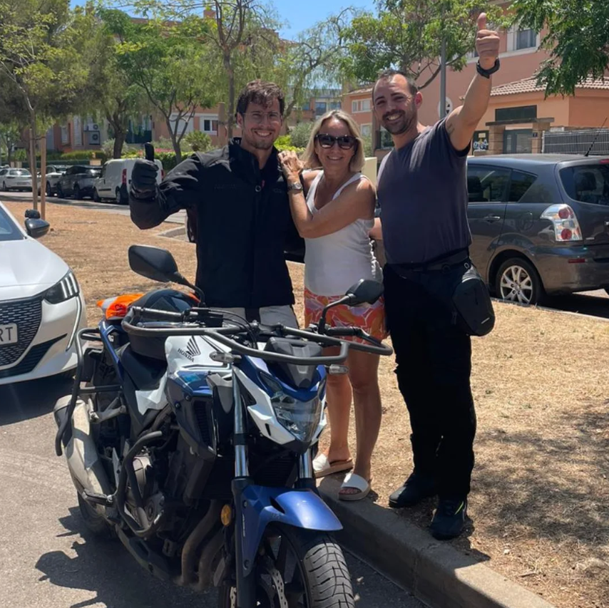 Tres personas sonrientes posando con una motocicleta en un parque, rodeadas de árboles y autos estacionados, en un día soleado.
