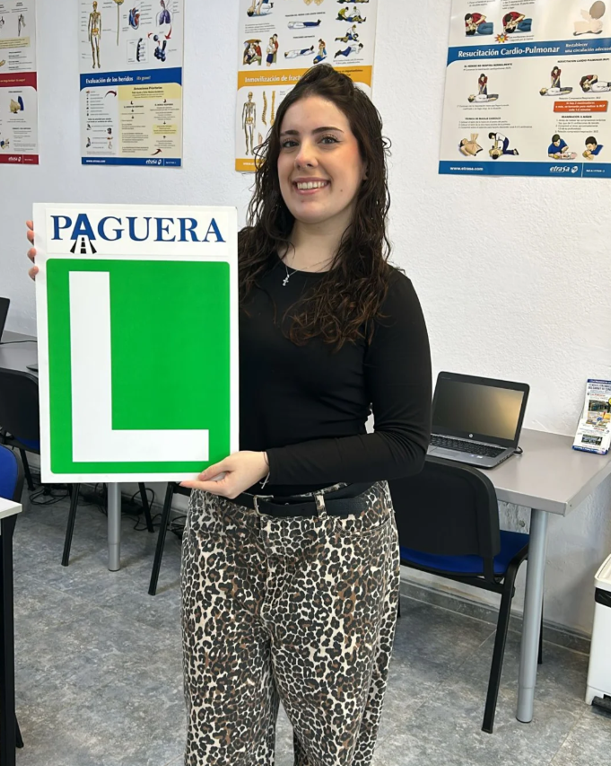 Mujer sonriendo sosteniendo señal de color verde con la letra L en un aula, en un entorno educativo con posters de primeros auxilios en la pared.
