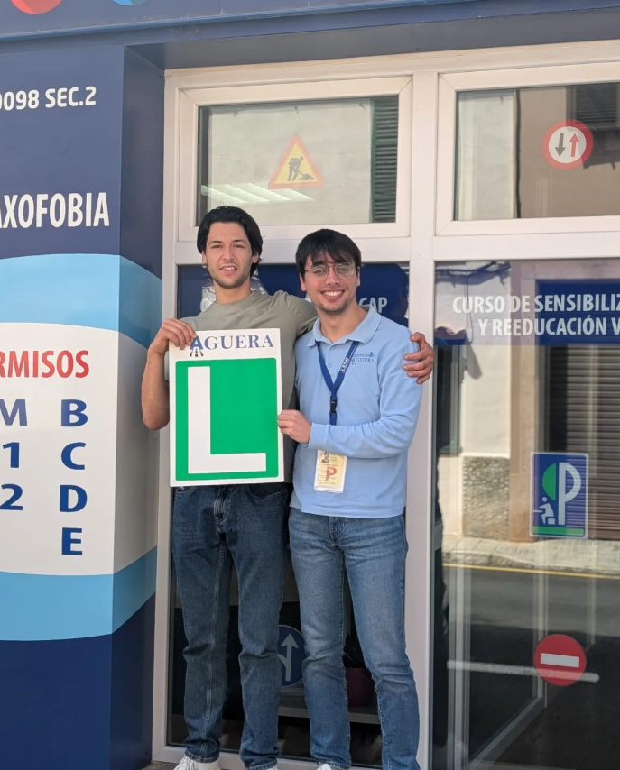 Dos jóvenes, uno con camiseta gris y otro con camisa azul, celebrando la obtención de su licencia de conductor nuevo junto a un instructor, en una oficina de autoescuela. Uno sostiene un cartel con una letra 'L'.