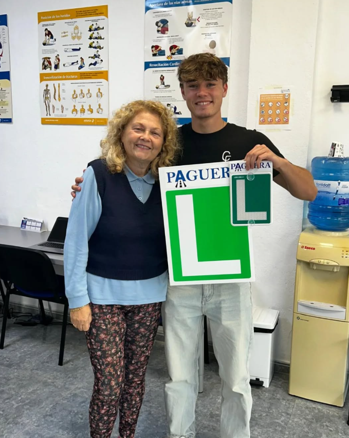 Joven y mujer en una oficina de tráfico, sosteniendo carteles de acompañamiento con letra L, con posters de primeros auxilios y señalización en la pared.