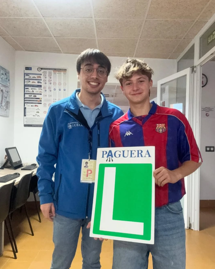 Un joven con camiseta del FC Barcelona sosteniendo un cartel verde con una 'L' y el nombre 'Paguera', acompañado por un hombre con uniforme azul y gafas en un consultorio o oficina.