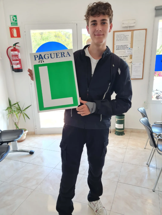 Joven sonriente sosteniendo una señal de aprendiz de conducir en una oficina.