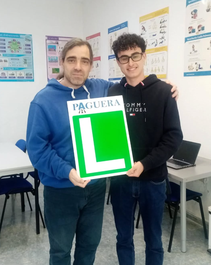 Dos personas, un adulto y un joven, sosteniendo un cartel con la letra 'L' y la palabra 'PAGUERA', en un aula con posters de educación en la pared.