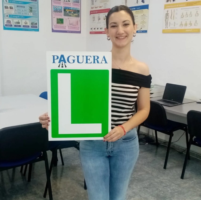Mujer sosteniendo una señal de carretera que dice 'PAGUERA' con una gran letra 'L' verde, en un aula con posters en la pared y computadoras en el fondo.