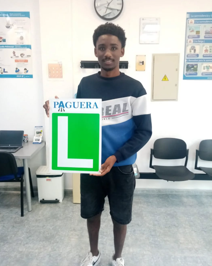 Joven sosteniendo cartel con la palabra "paguera" y una letra 'L' grande en color verde, en una sala de espera o institución educativa.