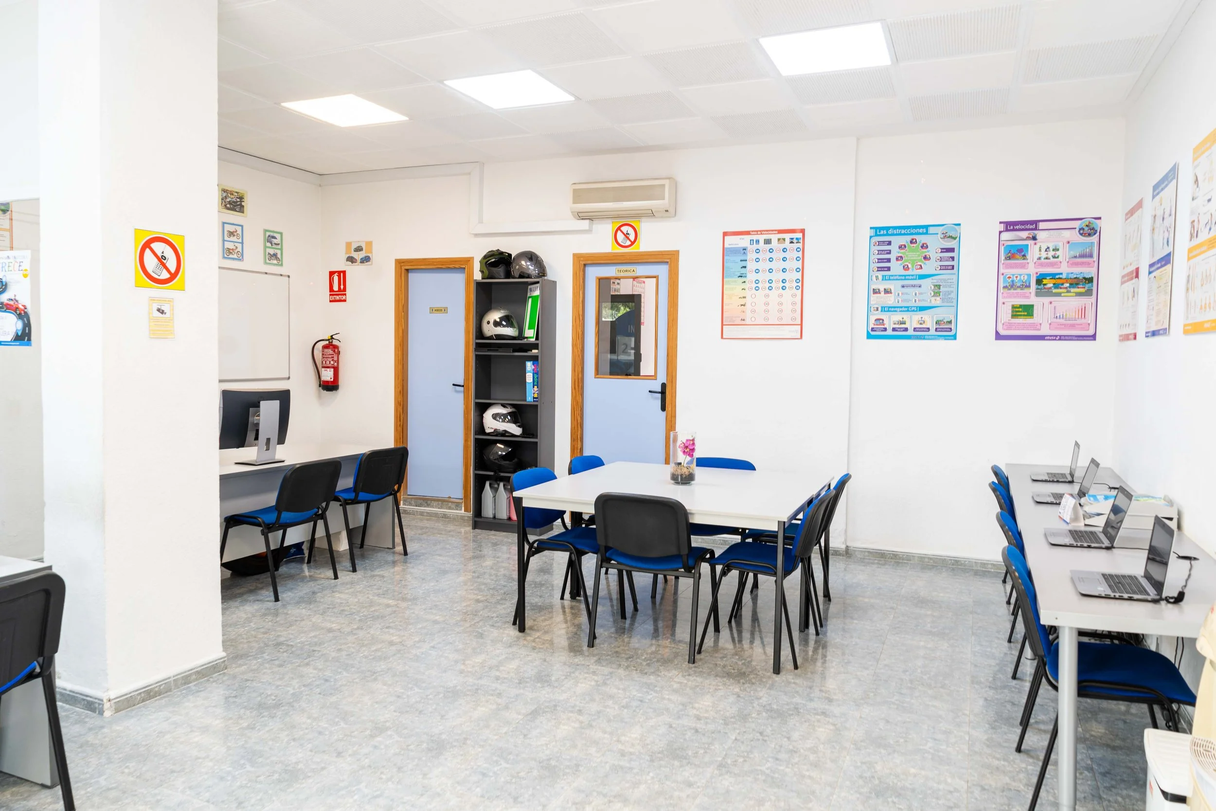 Sala de clases con mesas, sillas, computadoras portátiles, cartelera educativa, cascos de motocicleta en estantería y carteles informativos en las paredes.