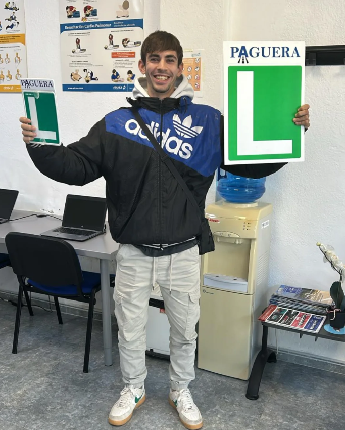 Joven sonriendo sosteniendo carteles de marca PAGUERA con la letra 'L', en una oficina con mesas, computadoras y un cartel de reanimación cardiopulmonar en la pared.