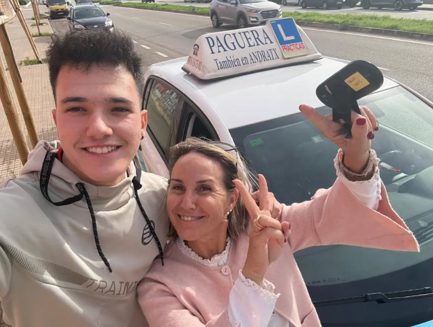 Dos personas, un joven y una mujer, posando con una coche de enseñanza de manejo, que tiene un cartel de 'PAGUERA' y una señal de 'L' para auto escuela en la calle, en un día soleado.